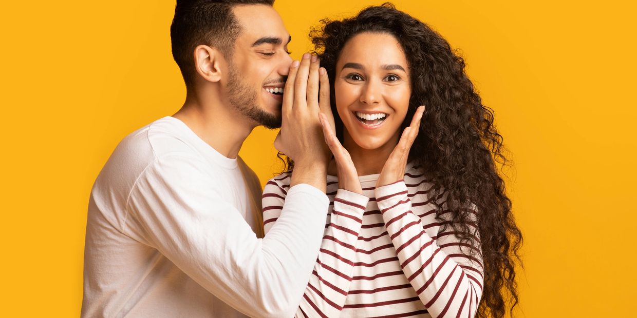 A man whispers a secret to a surprised woman against a yellow background.