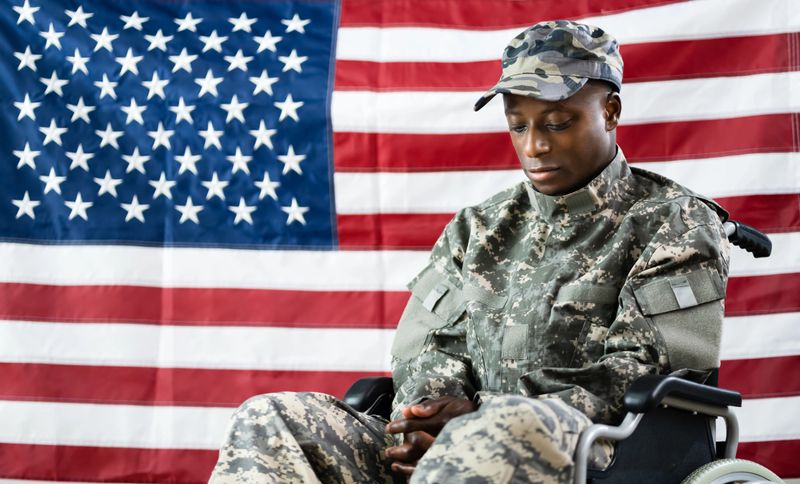 Patriotic Soldier Sitting In Wheel Chair Against American Flag