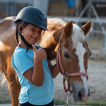 A young girl wearing a helmet gives a thumbs-up while standing next to a brown and white horse.