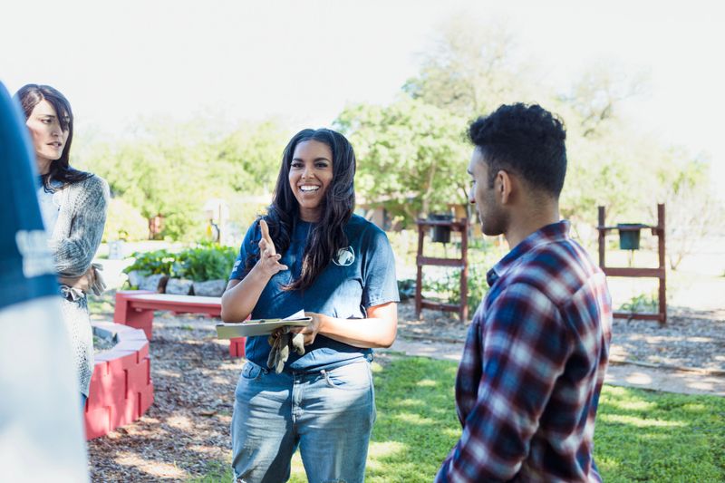 The mid adult female coordinator smiles and gestures as she trains a diverse group of volunteers at the community gardens.