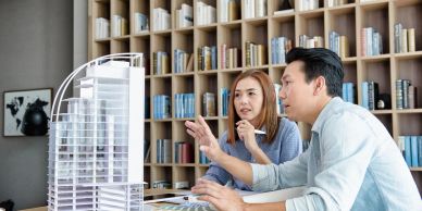 Two architects discussing a building model in an office with a bookshelf background.