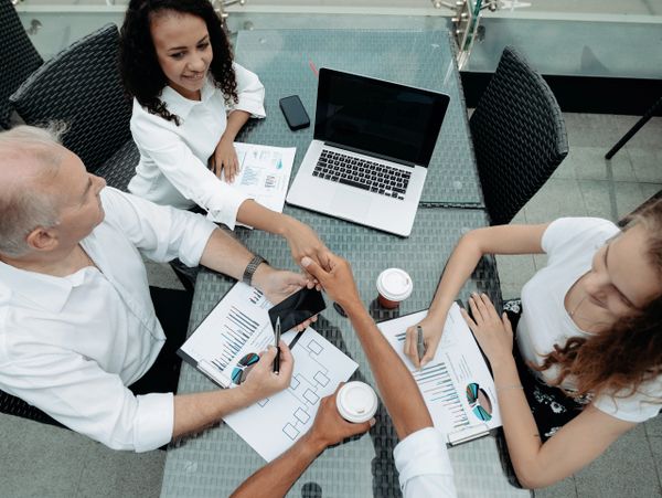 Four professionals discussing charts and shaking hands around a glass table outdoors.