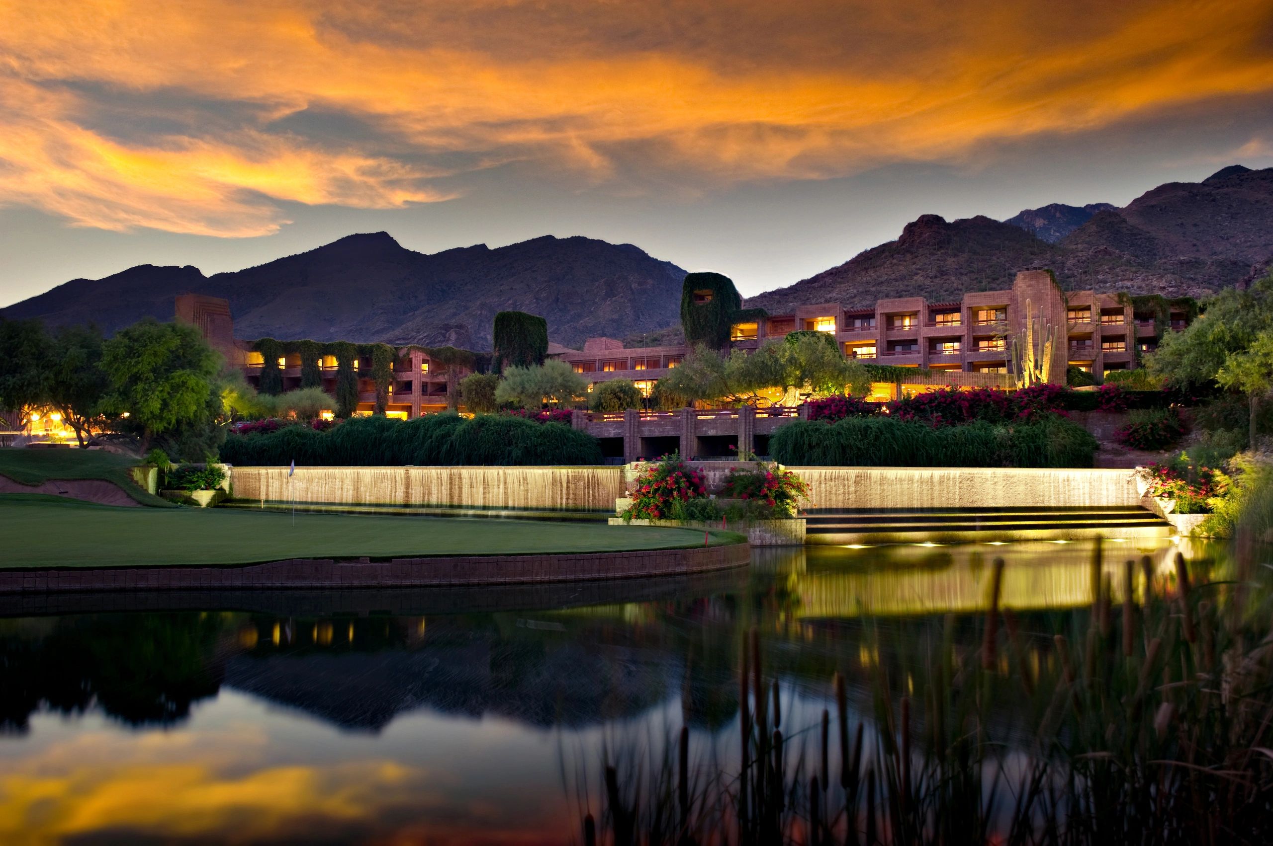 Resort at sunset with mountain backdrop and water feature.