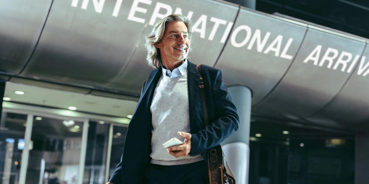 A man smiling and holding a phone at an international arrivals area.