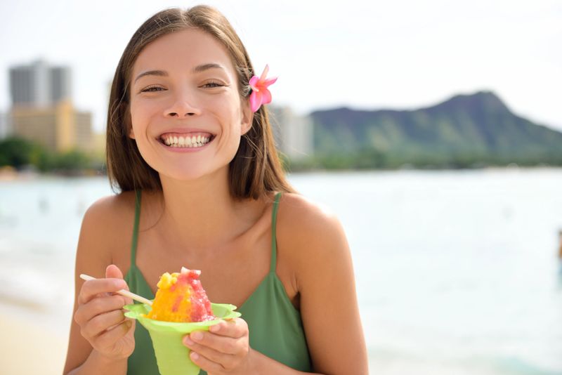 Hawaii woman on Waikiki Beach eating Hawaiian shave ice, a local shaved ice dessert. Happy smiling mixed race Asian Caucasian female model enjoying traditional Hawaiian snack. Oahu, Hawaii, USA.