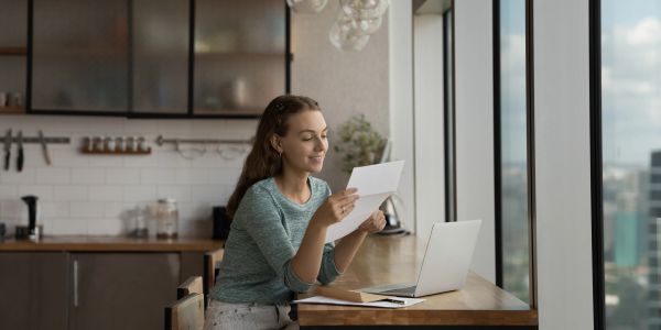 Woman reading a letter near a laptop by the window.