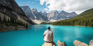 Person sitting on a rock overlooking a turquoise lake surrounded by mountains.