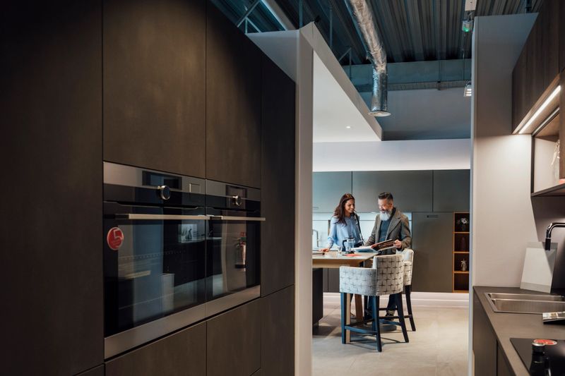 A wide-view shot of a mid-adult woman viewing a modern kitchen showroom interior with her father, she is considering buying a new kitchen to improve her home. They are reading through an in-store leaflet.