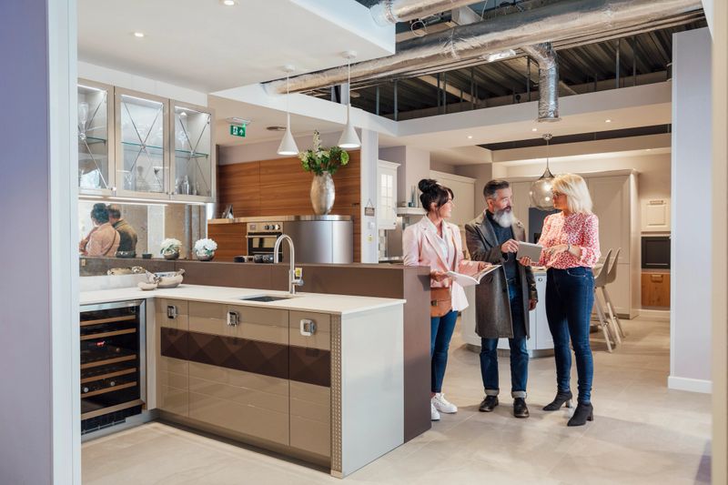 A wide-view shot of a mature couple viewing a modern kitchen showroom interior, they are considering buying a new kitchen to improve their home. A saleswoman is assisting the mature couple, she is holding a digital tablet.
