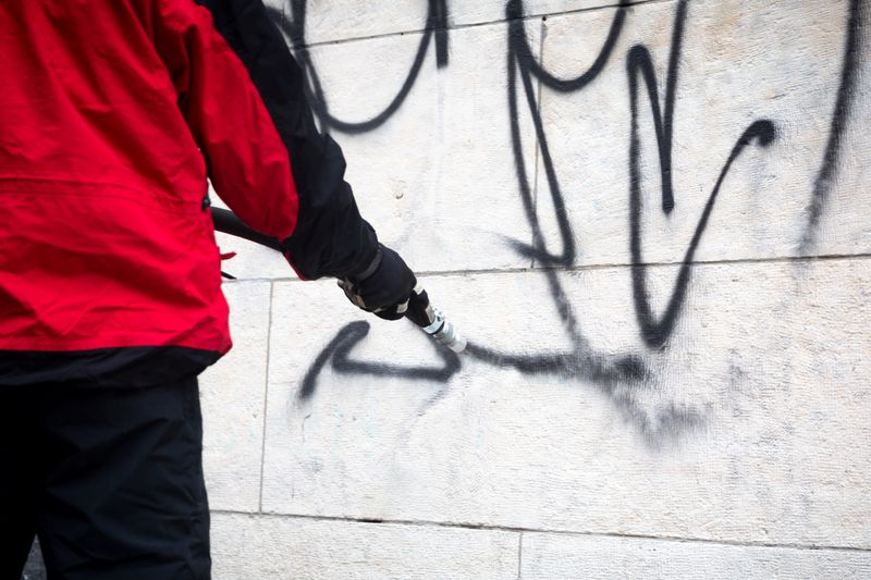 Worker cleans graffiti from a public building wall using pressure sand blaster.