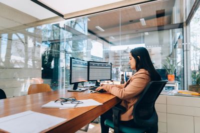 A woman working on dual monitors in a modern office with glass walls.