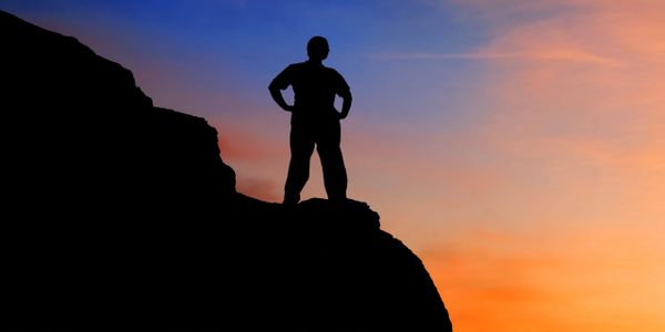 Silhouette of person standing triumphantly on a cliff at sunset.