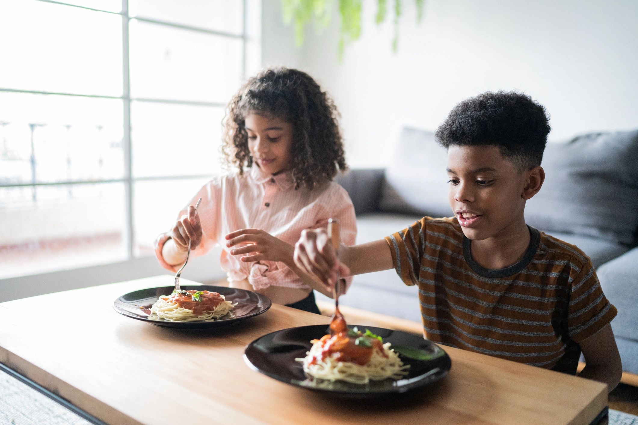 Two kids enjoying spaghetti with sauce at a bright dining table.