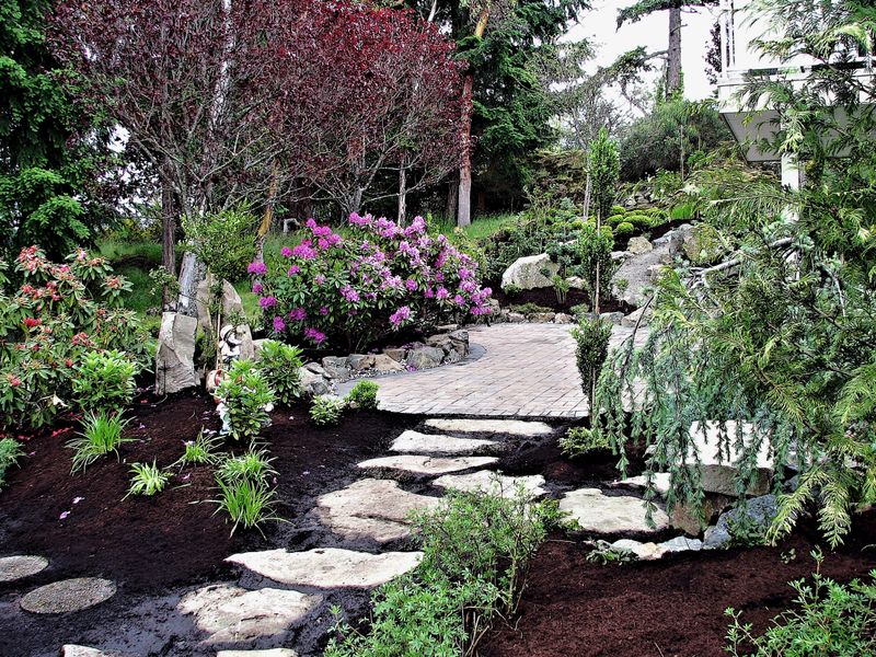 A beautiful footpath made of sandstone stepping stone leading through a formal garden landscape onto a paving stone deck surrounded by trees and plants in bloom.
