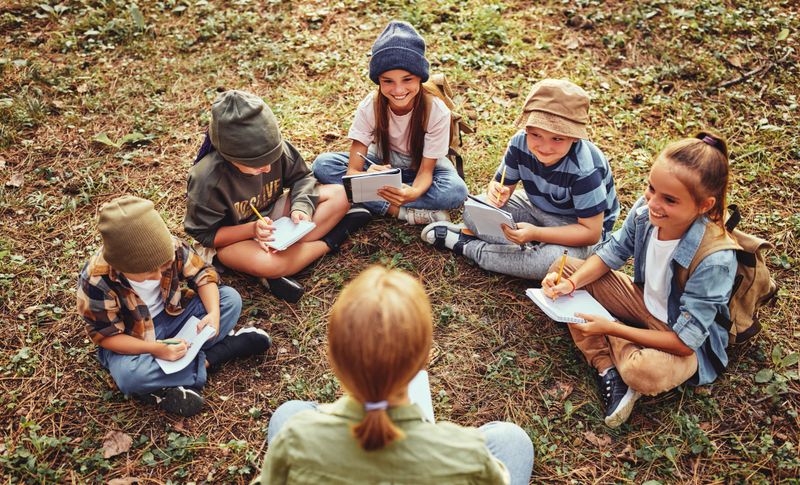 School activities in nature. Group of curious children and young female teacher sitting together on green grass in forest and learning about environment, boys and girls during ecology lesson in wood