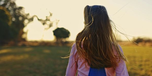 Girl with long hair stands outdoors at sunset, back to camera.