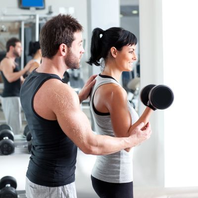 A man guides a woman lifting a dumbbell in a gym.