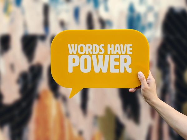 Hand holds a yellow speech bubble sign with the text 'WORDS HAVE POWER'.