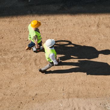 Two construction workers walking on dirt wearing helmets.
