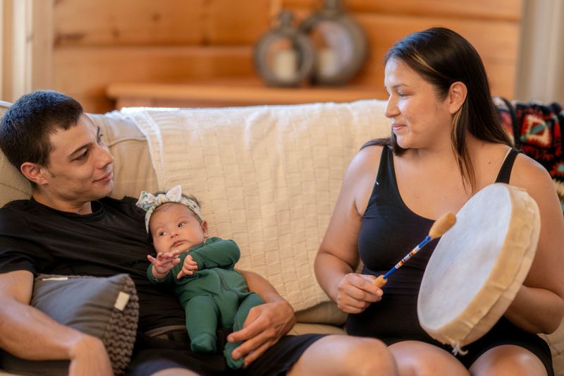 A beautiful young Indigenous family spending quality time together at home. The father is holding her newborn baby daughter. The mother is seated next to them and playing some music using traditional drums.