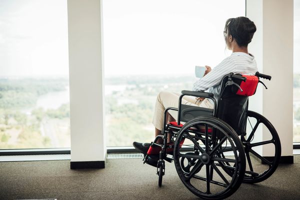 Woman in a wheelchair enjoying a drink while looking out a large window.