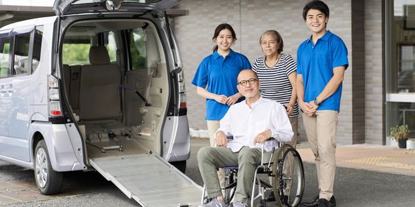 Elderly man in wheelchair with caregivers and a family member near accessible van ramp.