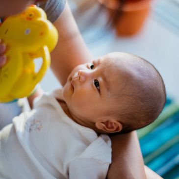 Baby gazing curiously at a yellow toy held by an adult.
