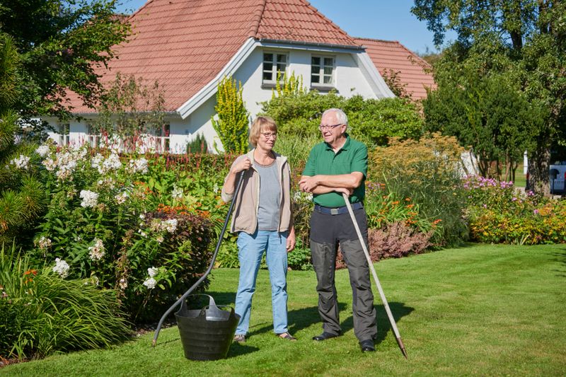 Standing on the lawn with gardening tool. Portraits of heterosexual couple in their backyard. Flowerbeds and bushes in the garden, One family detached house. Keeping in shape with gardening.