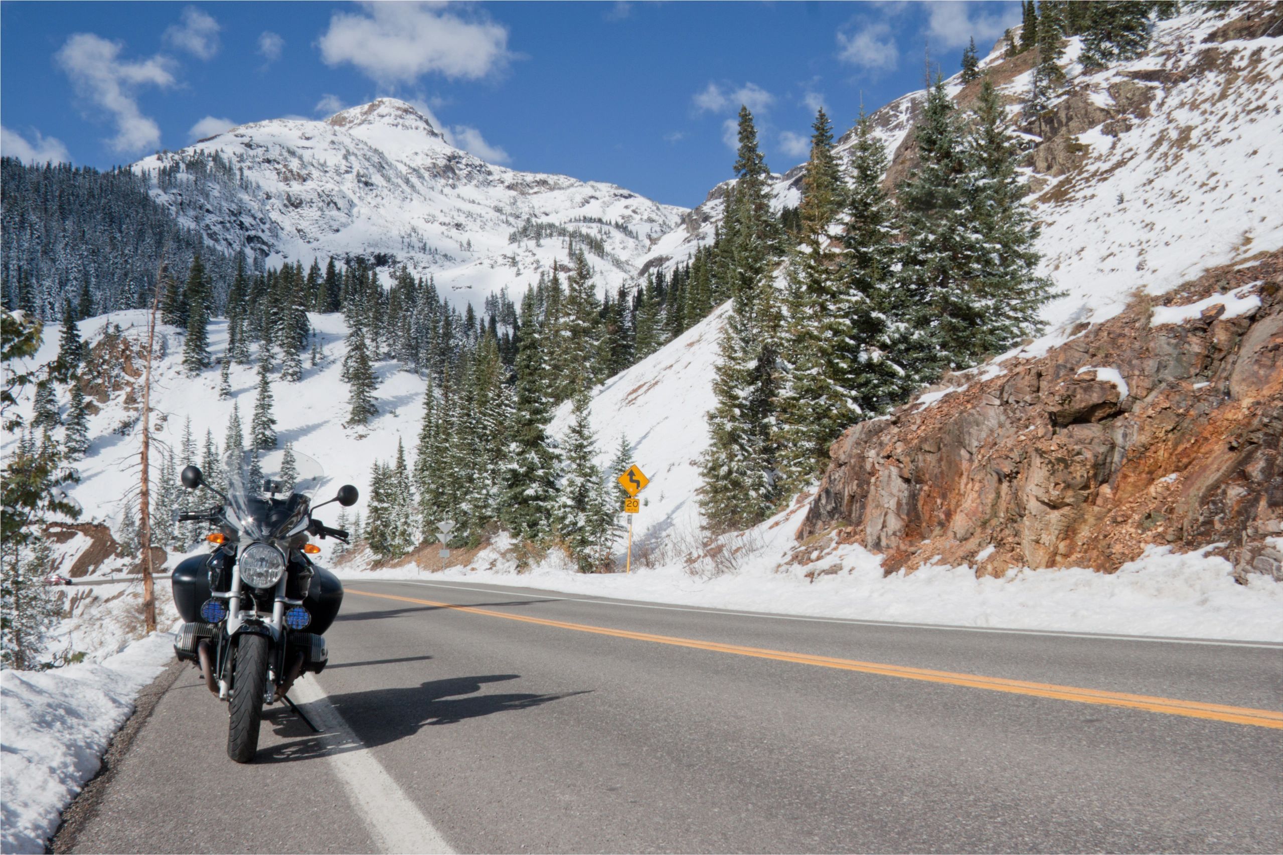 Motorcycle parked on a snowy mountain road with pine trees and blue sky.