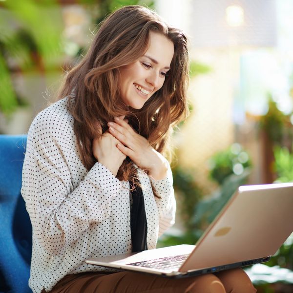 A joyful woman smiling at her laptop in a bright, plant-filled room.