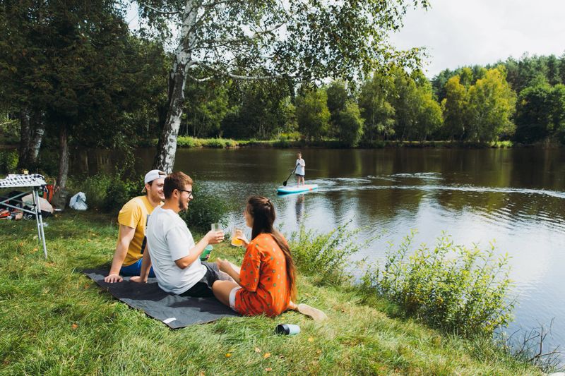 Young women and men sitting at the meadow near the lake with the picnic, talking, smiling, floating on the SUP paddle board and enjoying the summertime season in the forest
