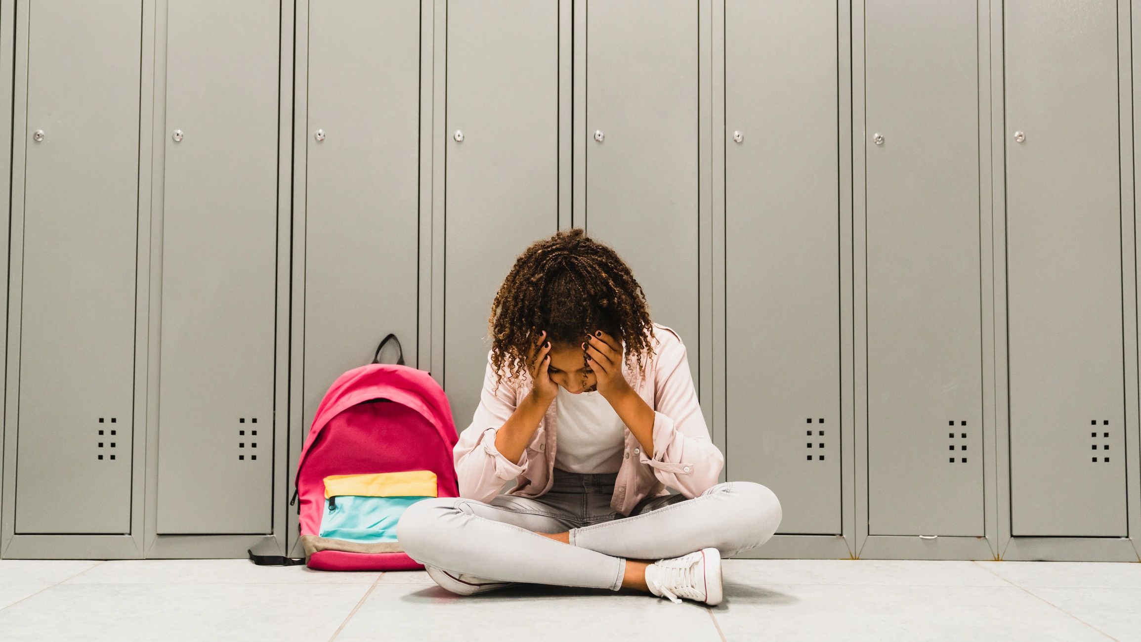 Girl sitting on floor cross-legged in front of school lockers with her head in her hands.