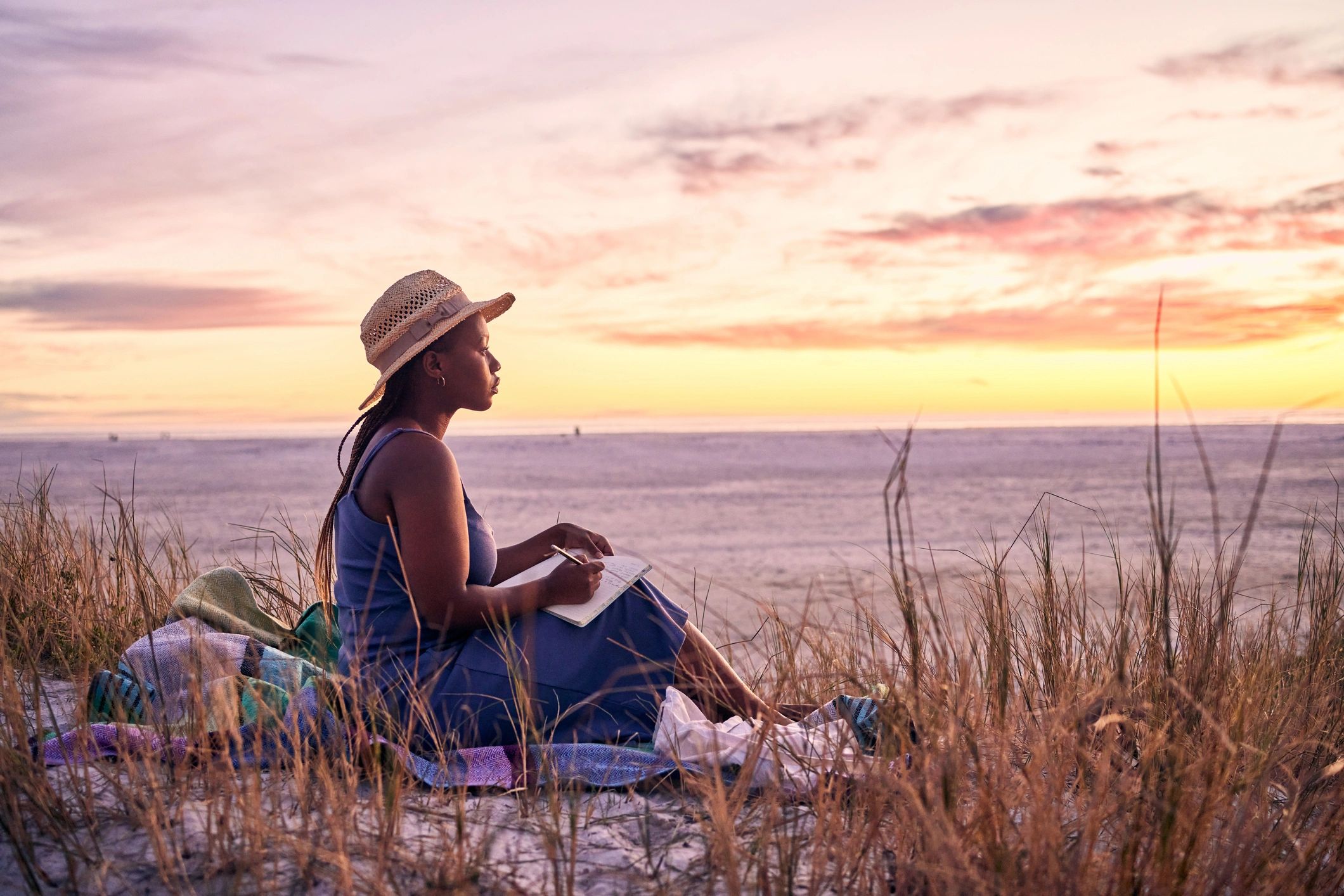 A woman in a hat writing in a notebook at sunset on the beach.