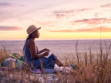 A woman in a hat writing in a notebook at sunset on the beach.