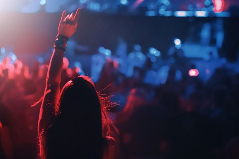 Closeup rear view of a late 20's woman dancing at a rave party. Lasers and spot lights firing in the background over big crowd after full year and a half of no public events.