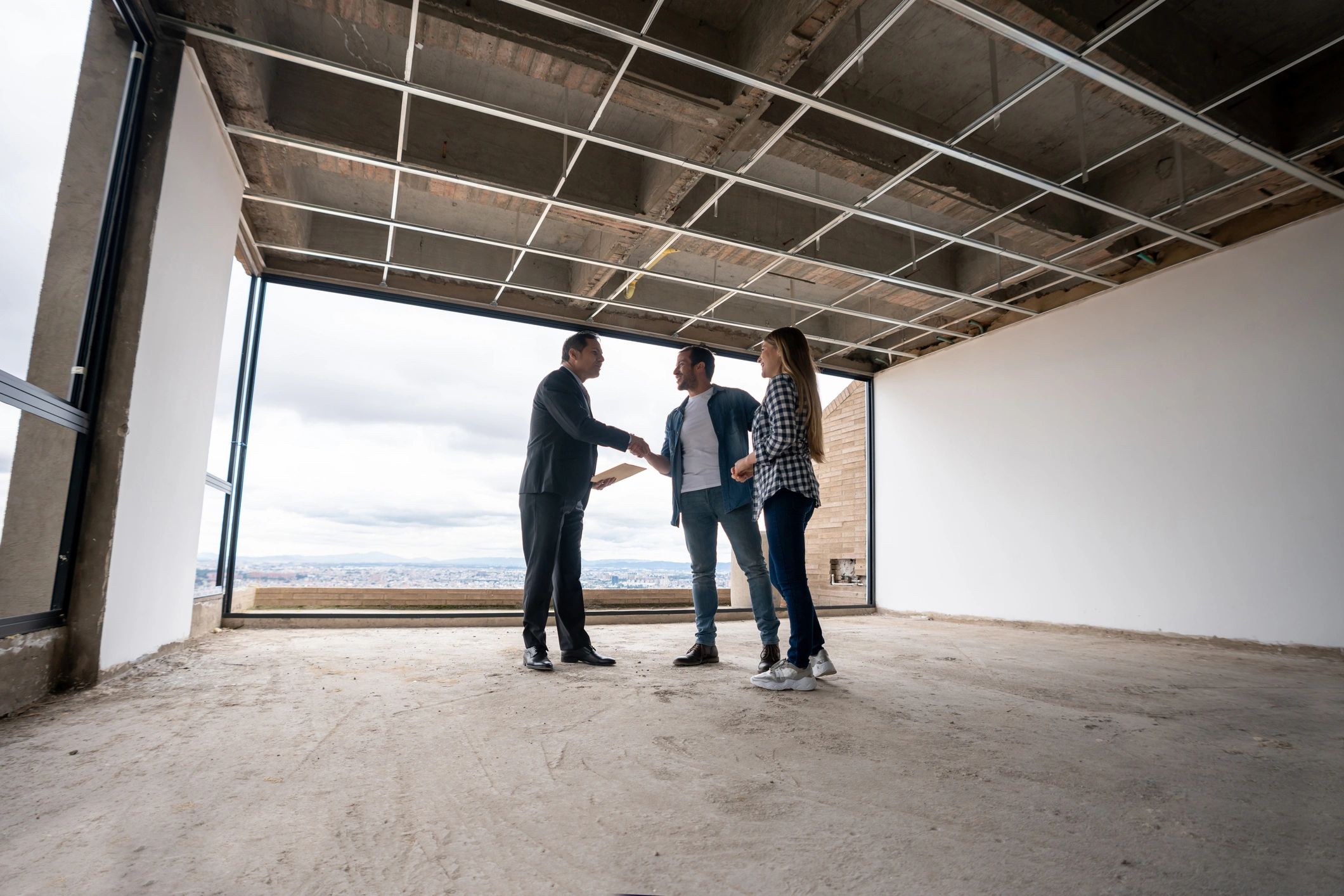 Couple shaking hands with a real estate agent in an unfinished apartment.