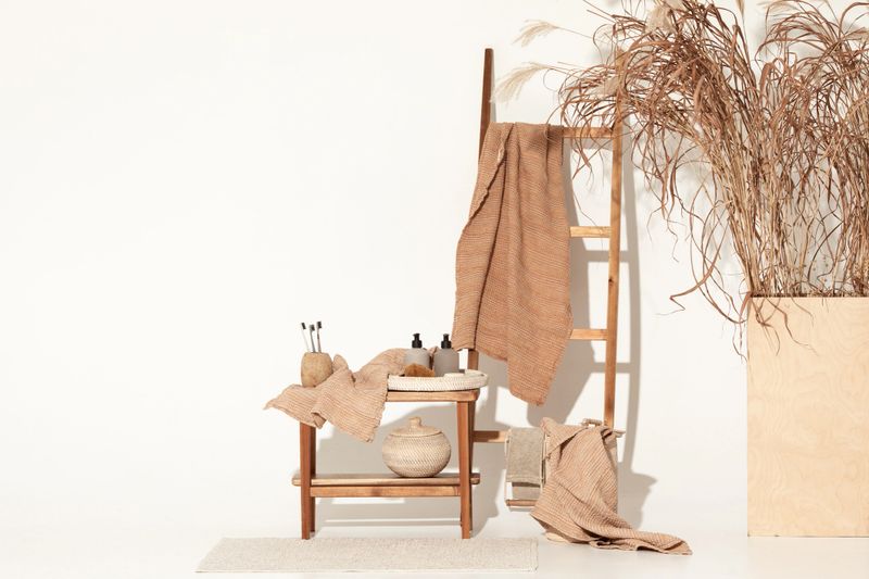 Natural waffle linen towels in earth tones on wood bench and towel ladder with bamboo toothbrushes, rattan baskets, and soap dispenser. Daily body care, spa and wellness zero waste bathroom concept