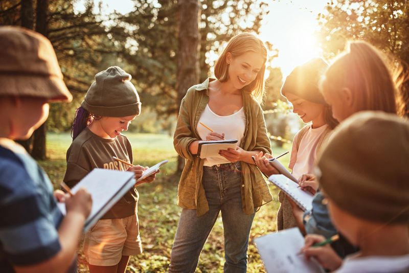 Young positive woman teacher interacting with group of school kids boys and girls during ecology lesson outdoors in forest, kids with notebooks learning about nature and wildlife on sunny autumn day