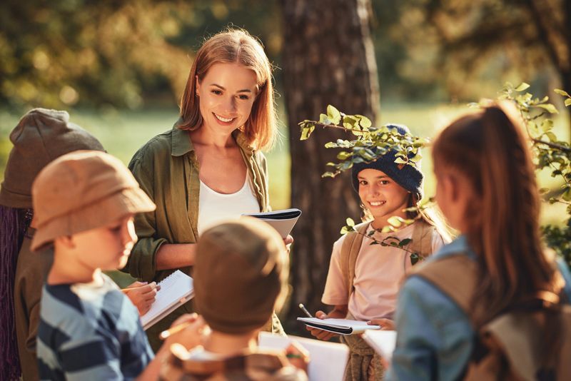 Group of curious school children with notebooks listening to their young female teacher while learning about nature together, looking at green leaf during ecology lesson in autumn forest on sunny day