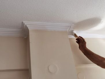 A close-up of a person's hand painting a white, ornate ceiling cornice with a brush.
