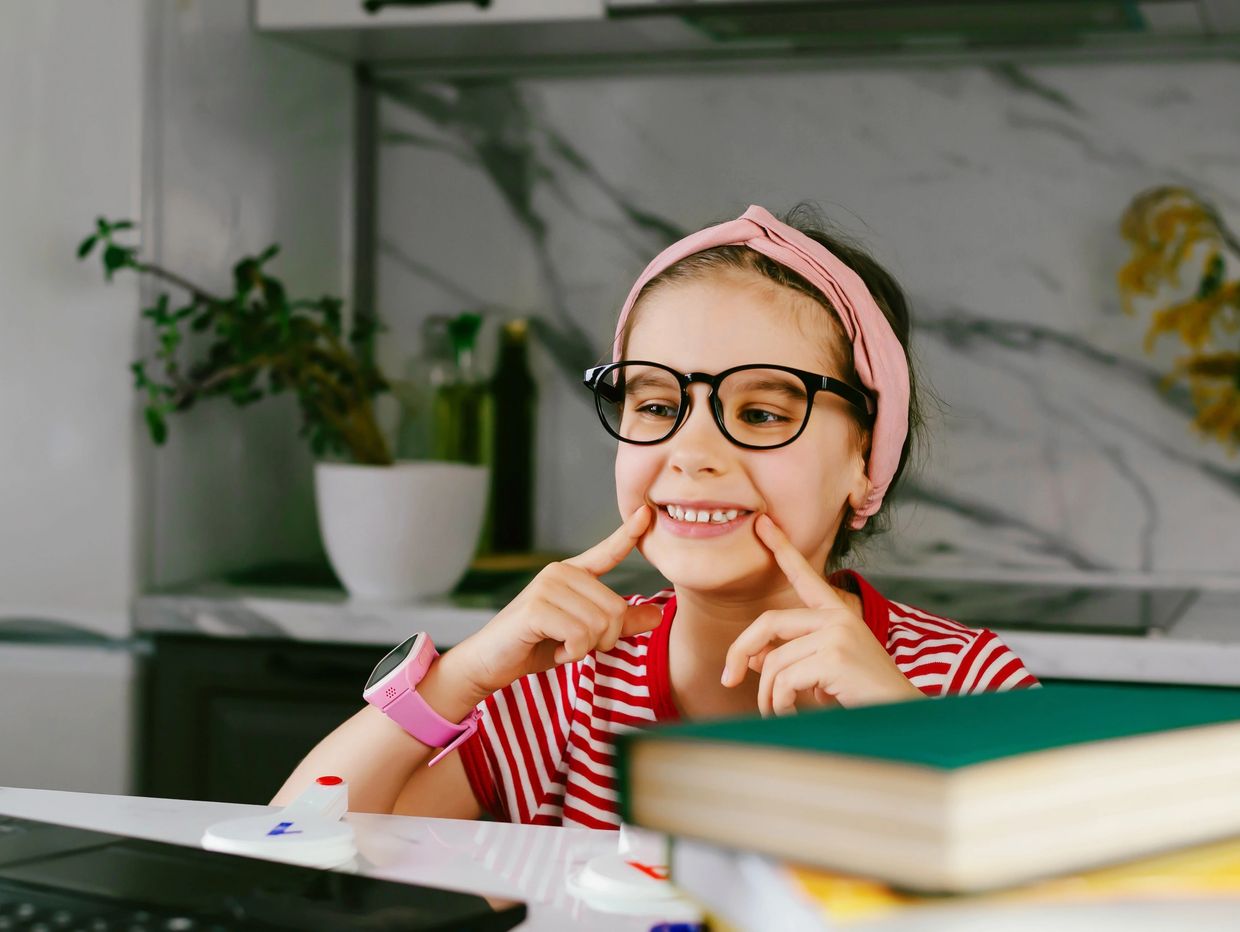 Young girl with glasses and a pink headband smiling at a laptop.