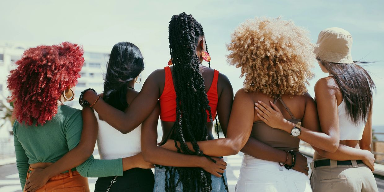 Five diverse women stand arm in arm, showing unity and friendship from behind.