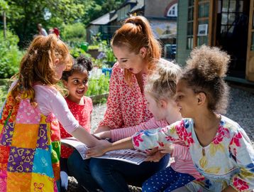 A woman and four children happily play a game outdoors, placing their hands together.