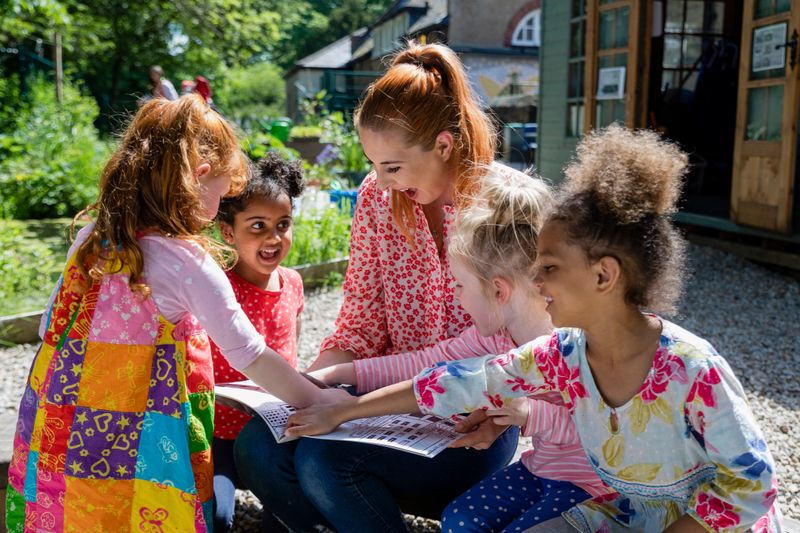 A front view of a teacher and her excited little pupils sitting and doing storytime outdoors in the schoolyard on the benches on a wonderful summer's day in Hexham in the North East of England.