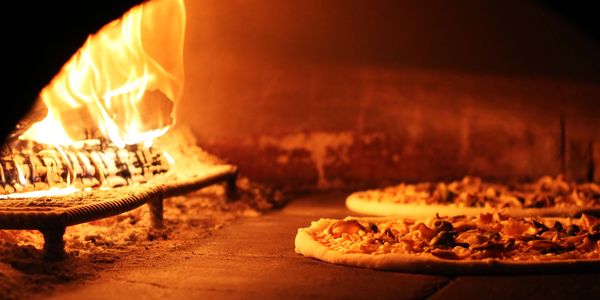 Two pizzas baking inside a wood-fired oven with a roaring fire.