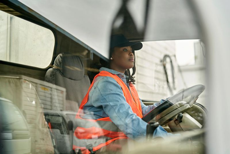Personal perspective of mid 20s woman in casual clothing, cap, and reflective vest sitting in driver’s seat, looking straight ahead, ready to begin road trip.
