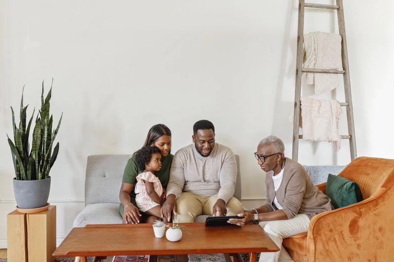 A mature adult businesswoman is having a meeting with a mixed race couple in their home. The husband and wife brought their toddler daughter to the casual meeting. The child is sitting on her mother's lap. The sales representative is using a tablet computer to help explain paperwork. The family is preparing for the future.