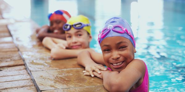 Three children in swim caps smiling by the pool edge.
