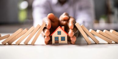 Hands protecting a small house model from falling dominoes.