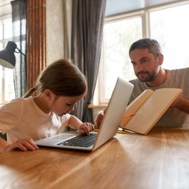 A young girl uses a laptop while an adult holds a notebook nearby.